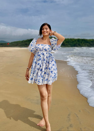 Woman in a white dress with blue floral pattern standing on a beach with ocean and sky in the background