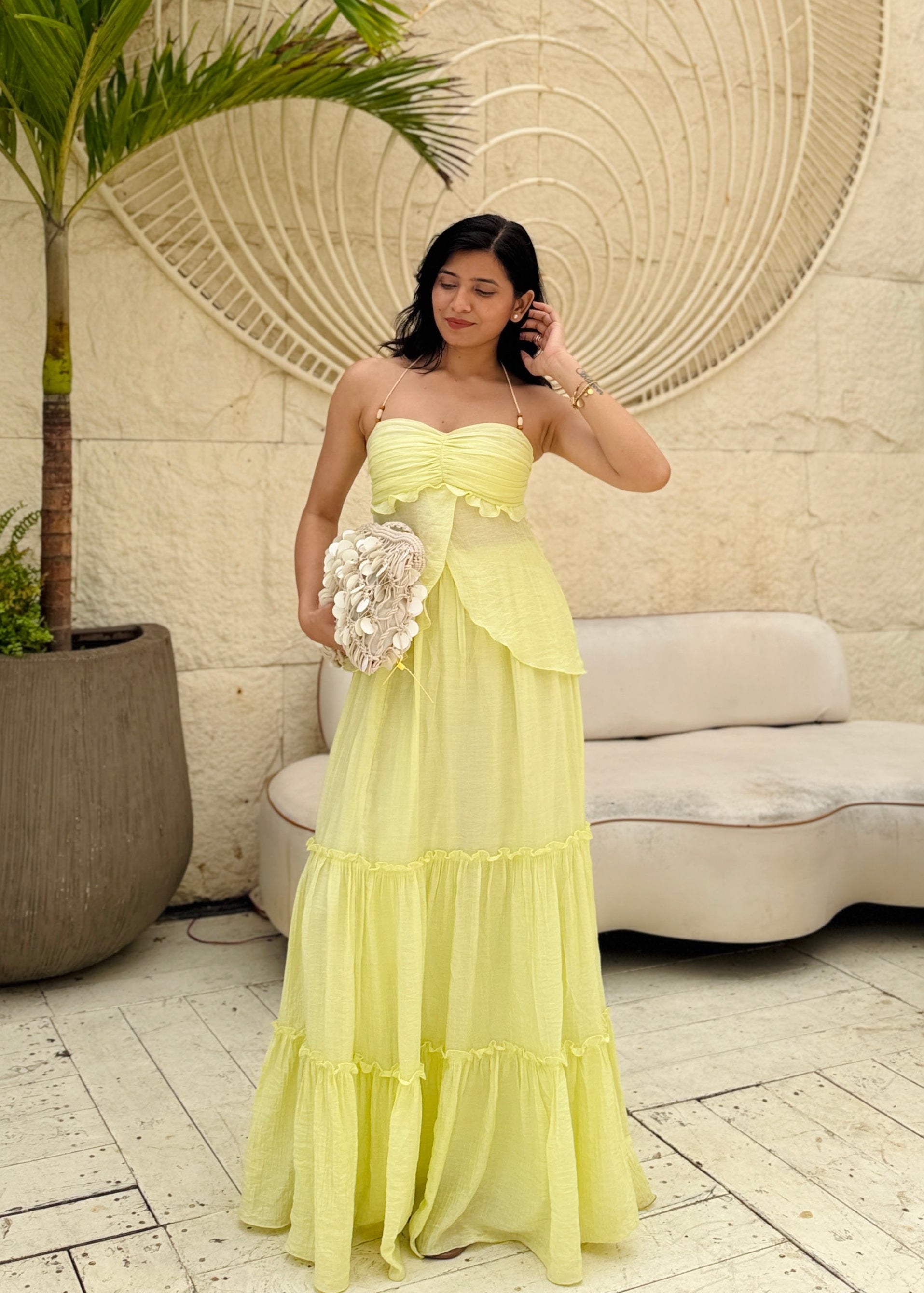 Woman in a yellow dress standing in front of a decorative wall with a palm tree and sofa in the background.