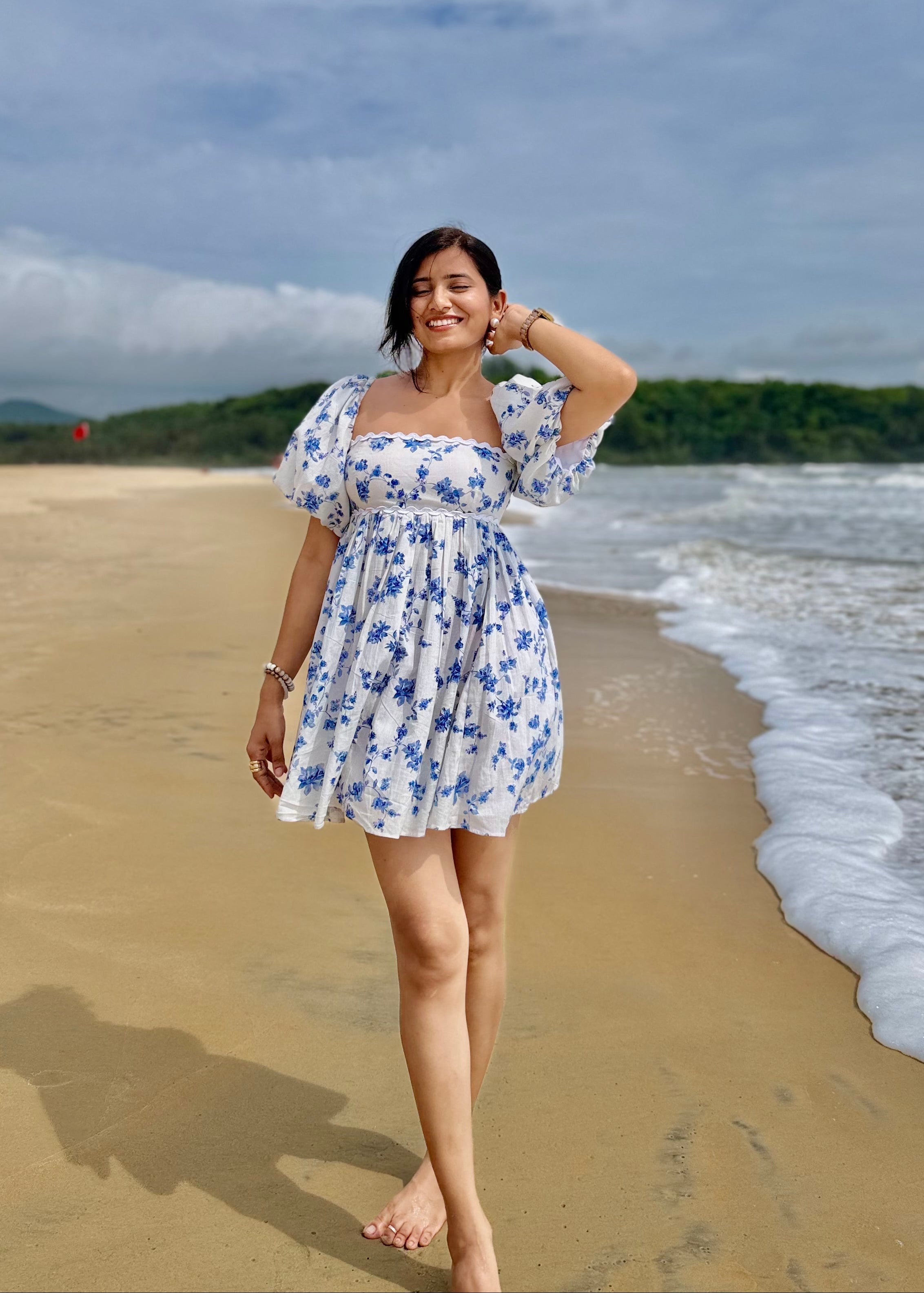 Woman in a white dress with blue floral pattern standing on a beach with ocean and sky in the background