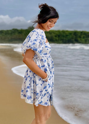 Woman in a floral dress standing on a beach with waves and greenery in the background