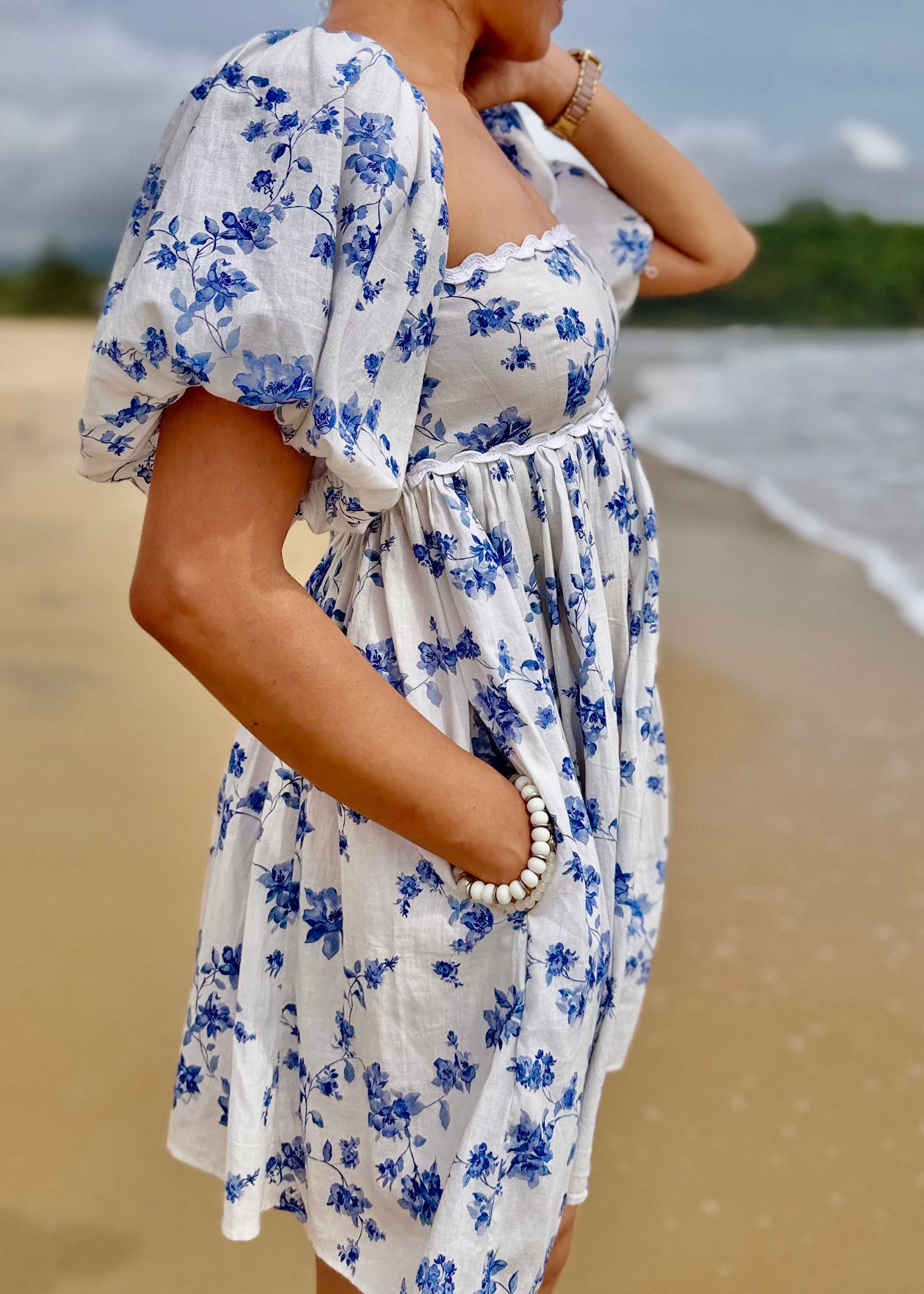 Woman in a white dress with blue floral pattern standing on a beach.