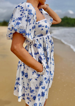 Woman in a white dress with blue floral pattern standing on a beach.