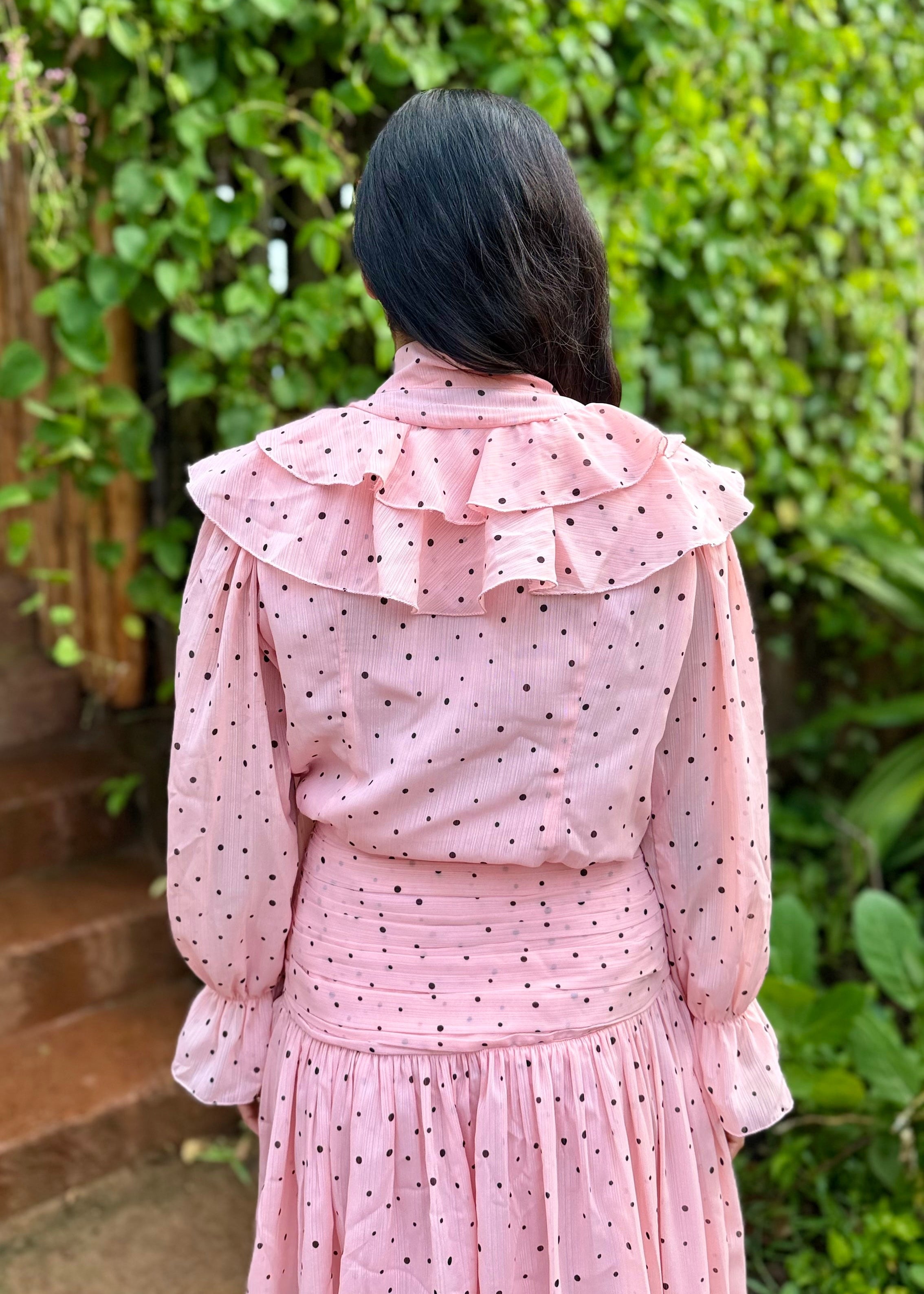 Person wearing a pink polka dot dress with a ruffled collar in front of green foliage.